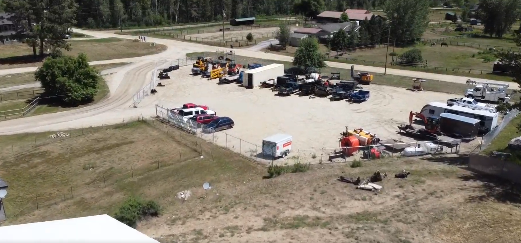 The fenced laydown yard at Bitterroot Industrial Park, holding skid steers, dump trailers, service trucks, cable spools, and equipment.